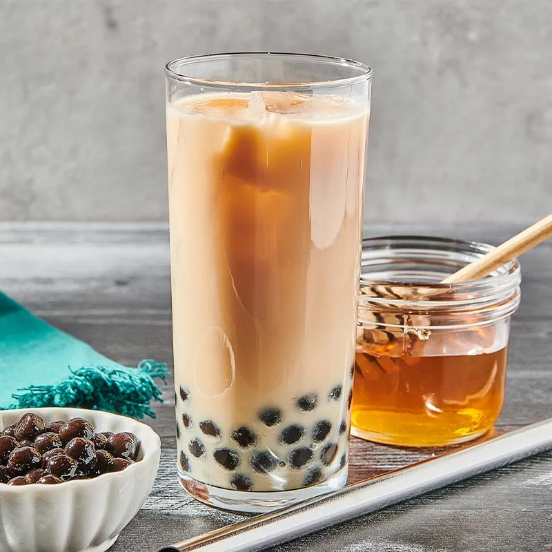 : Verre de thé au lait perlé avec perles de tapioca noires, accompagné d'un pot de miel et d'une cuillère en bois, sur une table en bois.